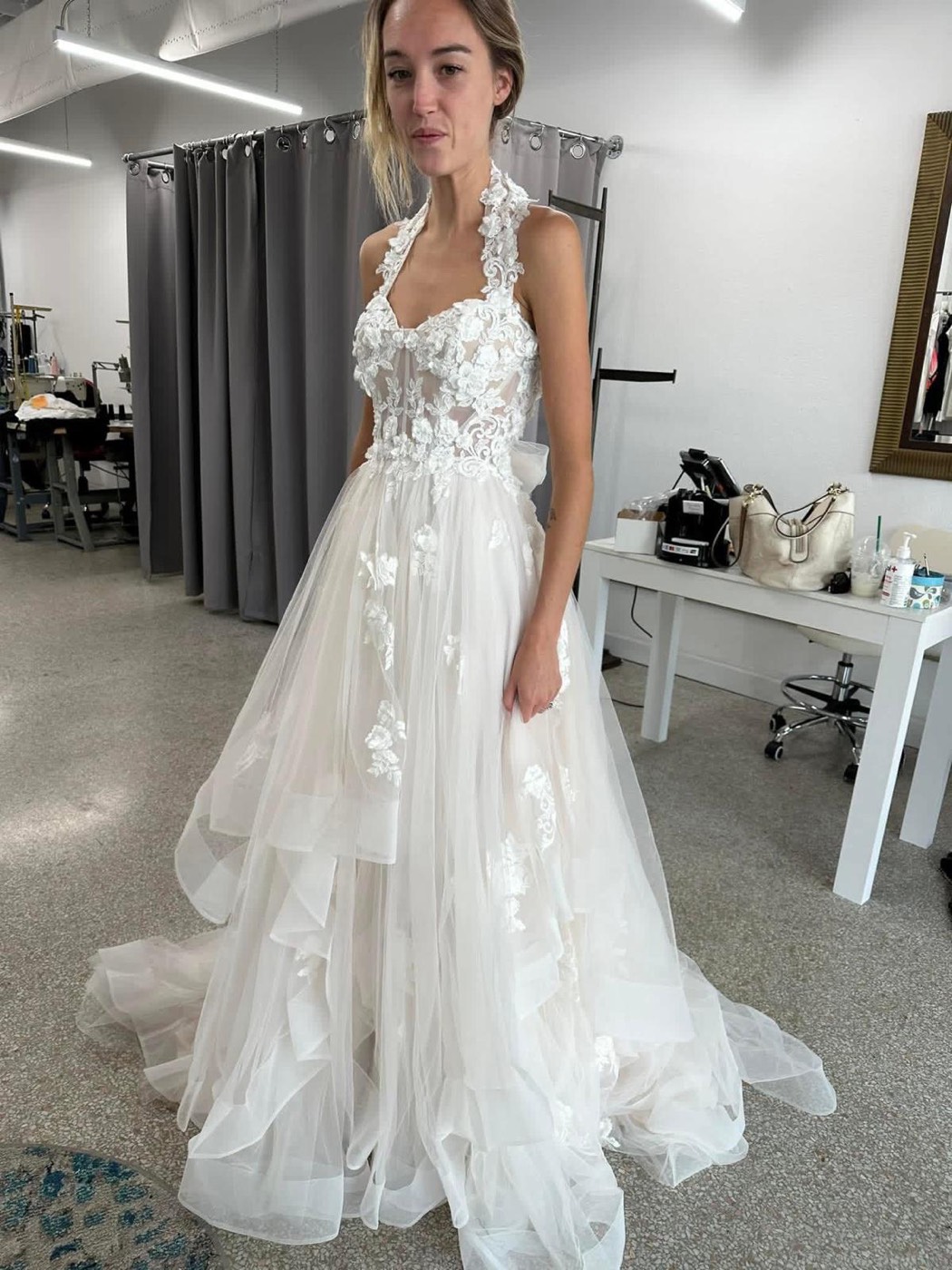 Bride in a halter-neck gown with a lace bodice and a layered tulle skirt, in front of the gray fitting-room curtain