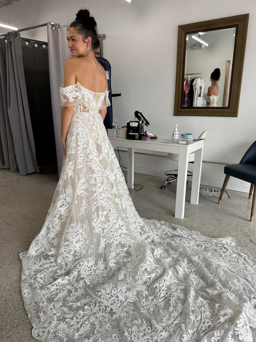Bride in an off-the-shoulder lace gown with a long train, seen from behind next to the fitting mirror