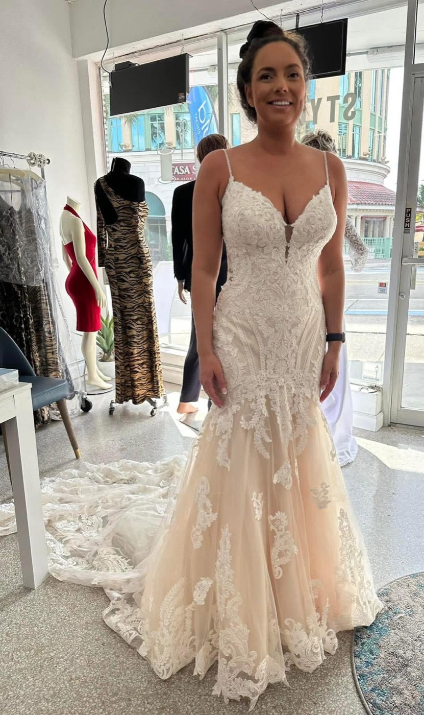 Bride in a fitted lace gown with thin straps and a blush skirt with a long train, by the shop window