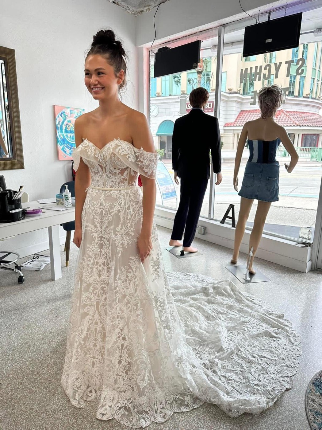Bride smiling in an off-the-shoulder lace wedding gown with a long train, standing by the shop window