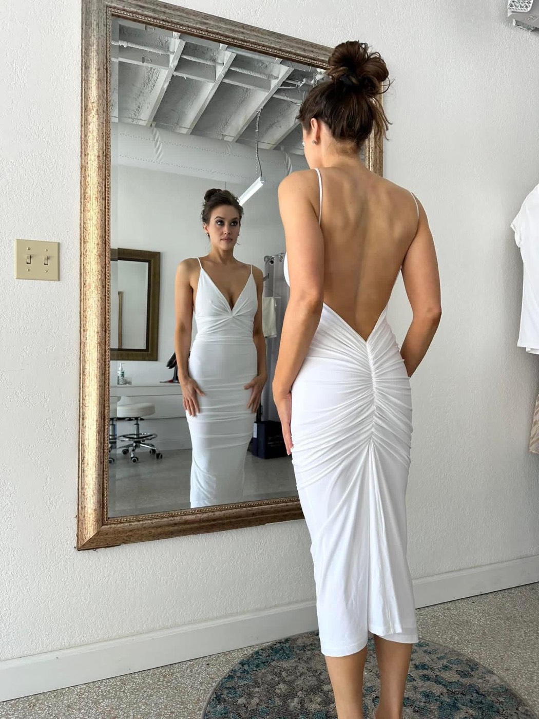 Back of a white ruched midi dress with an open back and thin straps, seen in a gold-framed mirror