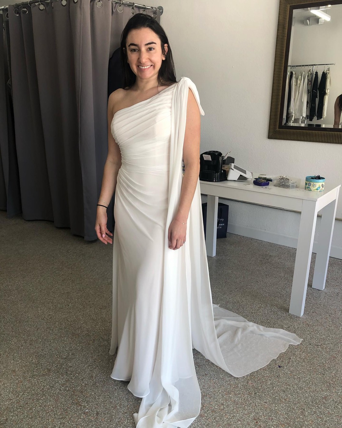 Woman in a white one-shoulder gown with a ruched bodice and a long chiffon sash that falls into a train, in front of the fitting-room curtain